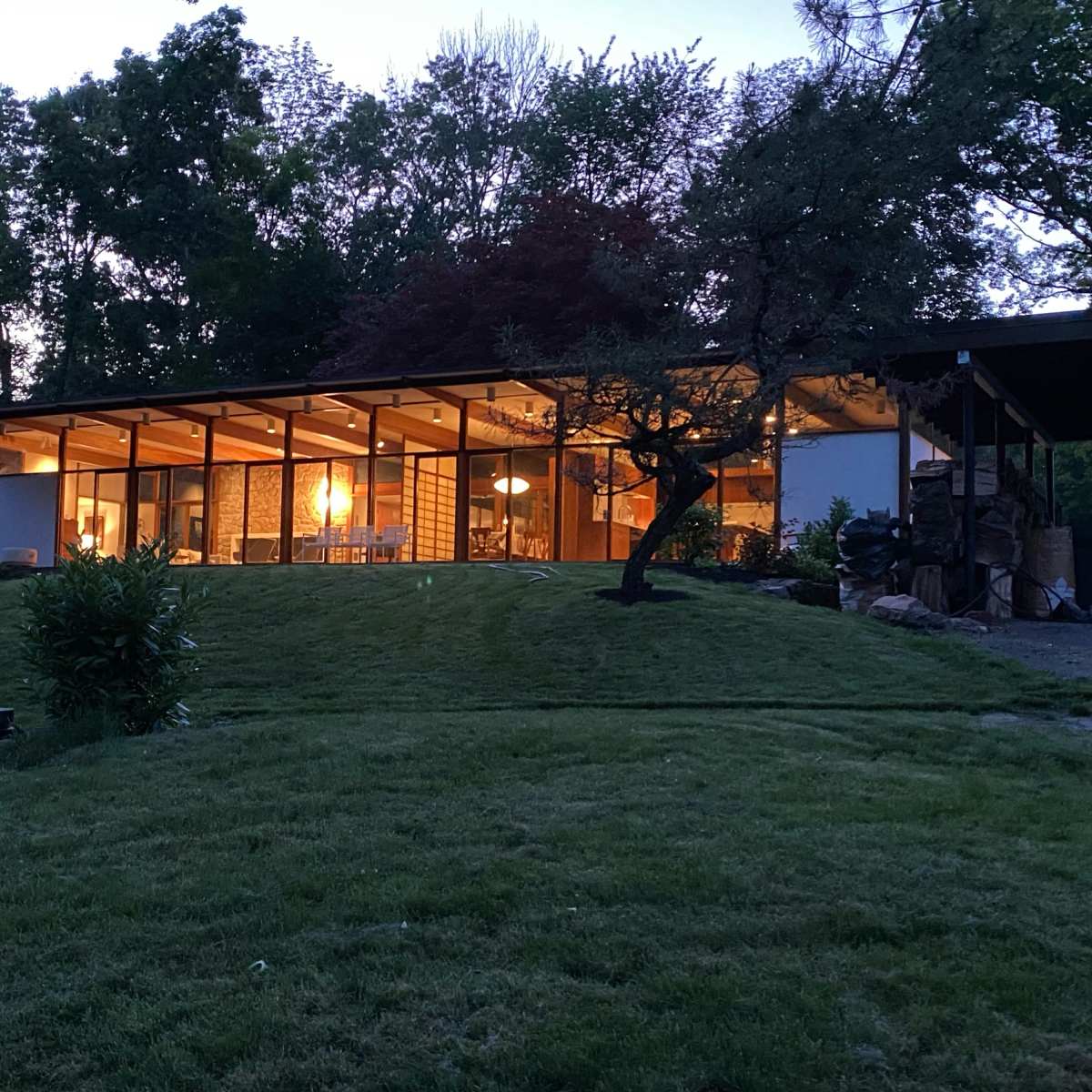 A modern glass and wood structure sits on a grassy hill, illuminated from within as dusk falls.