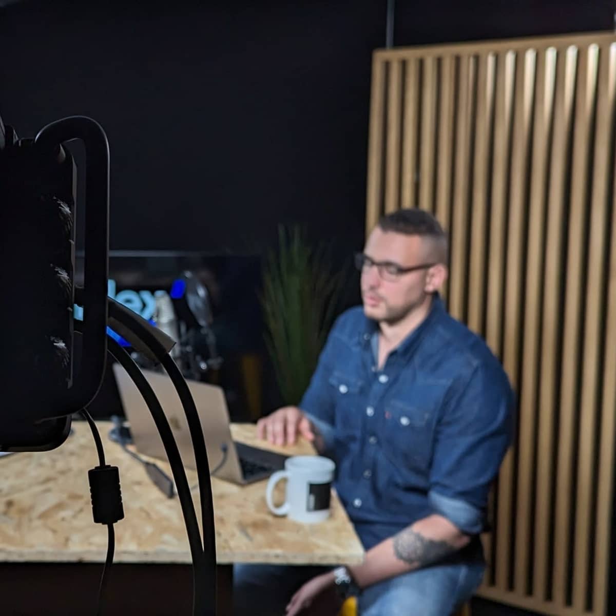 A video recording setup shows a man in a denim shirt seated at a table with a coffee mug, while another man is partially visible on a screen in front of a backdrop with wooden panels.