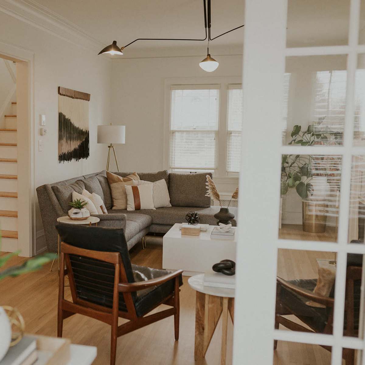 A cozy living room with a gray couch, two wooden chairs, and a staircase in the background.