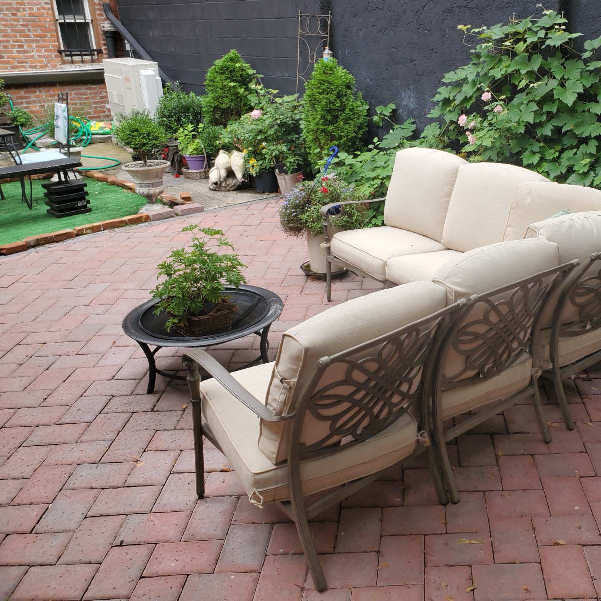 A patio area with a beige sofa set and armchairs arranged around a circular black firepit, surrounded by greenery and brick paving.