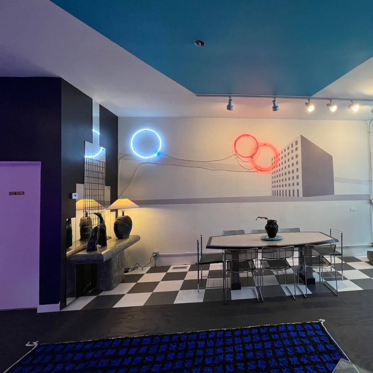 A modern interior space featuring a dining area with a black and white checkered floor, a table and chairs, and colorful neon wall art.