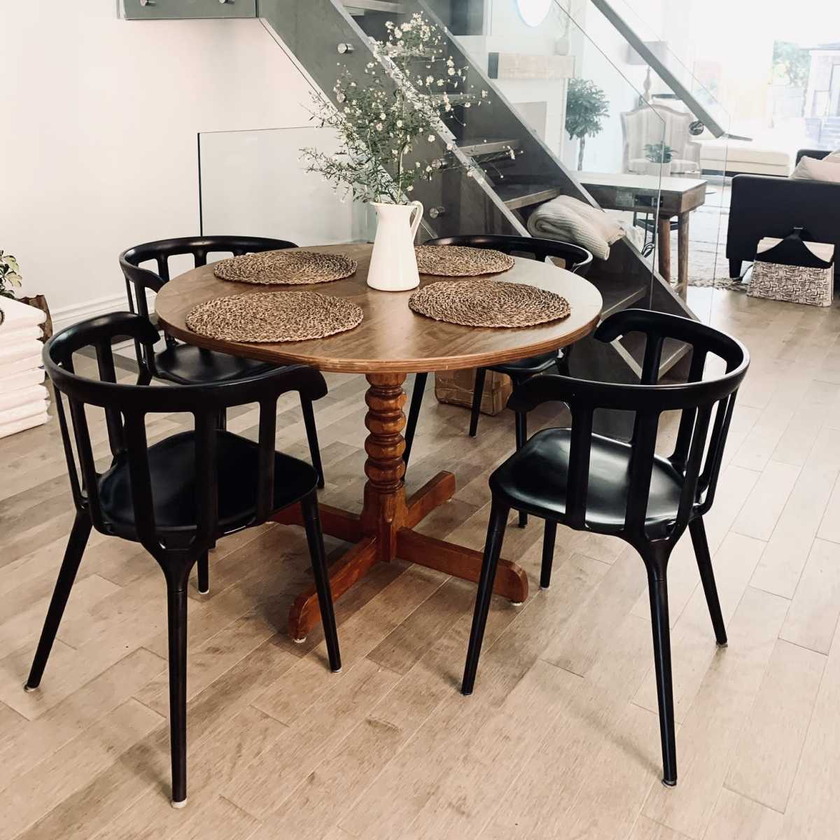 A round wooden dining table with four black chairs is arranged beneath a staircase with glass railing in a modern interior space.