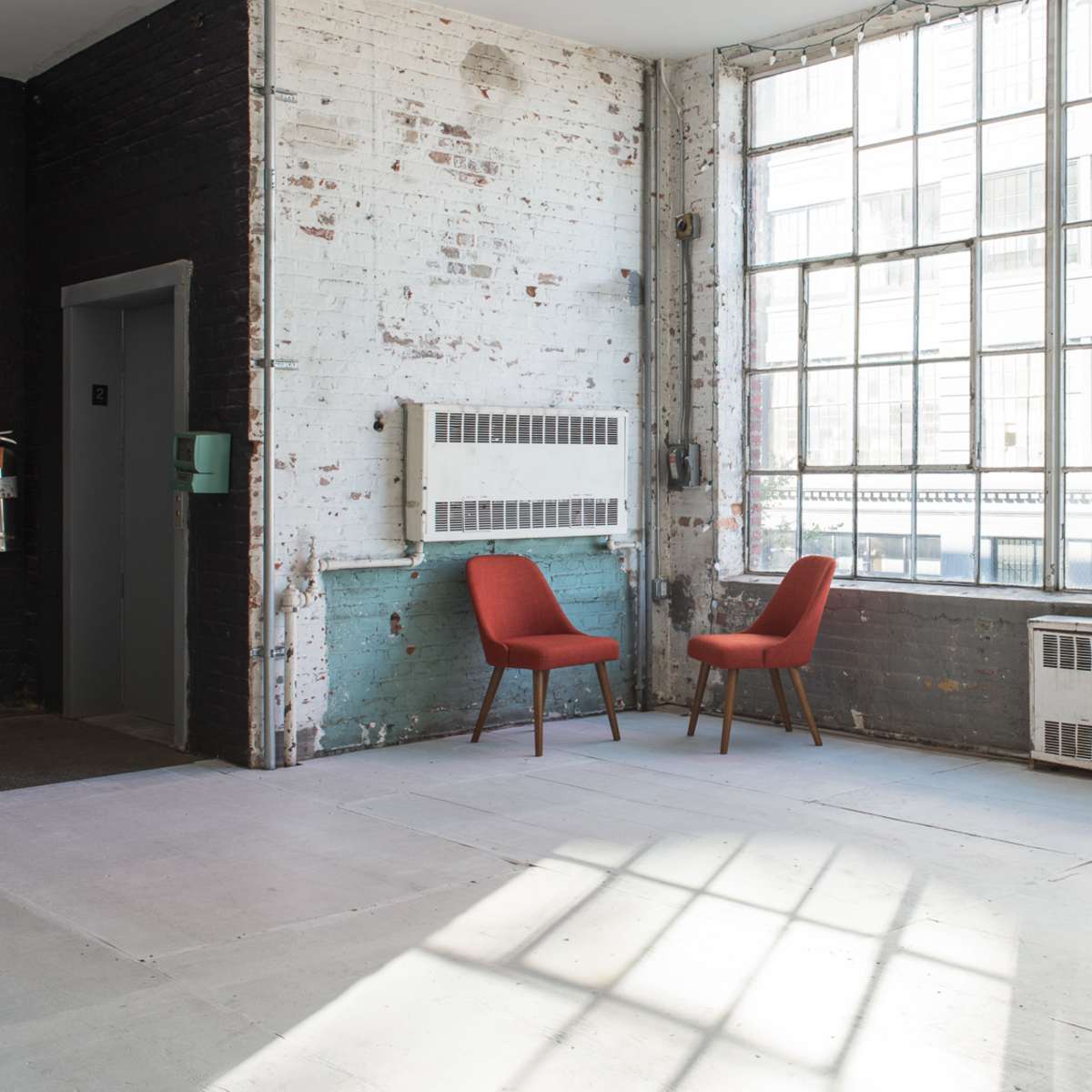 A spacious industrial room with two red chairs positioned near a large window, and a white shelving unit standing against the wall.