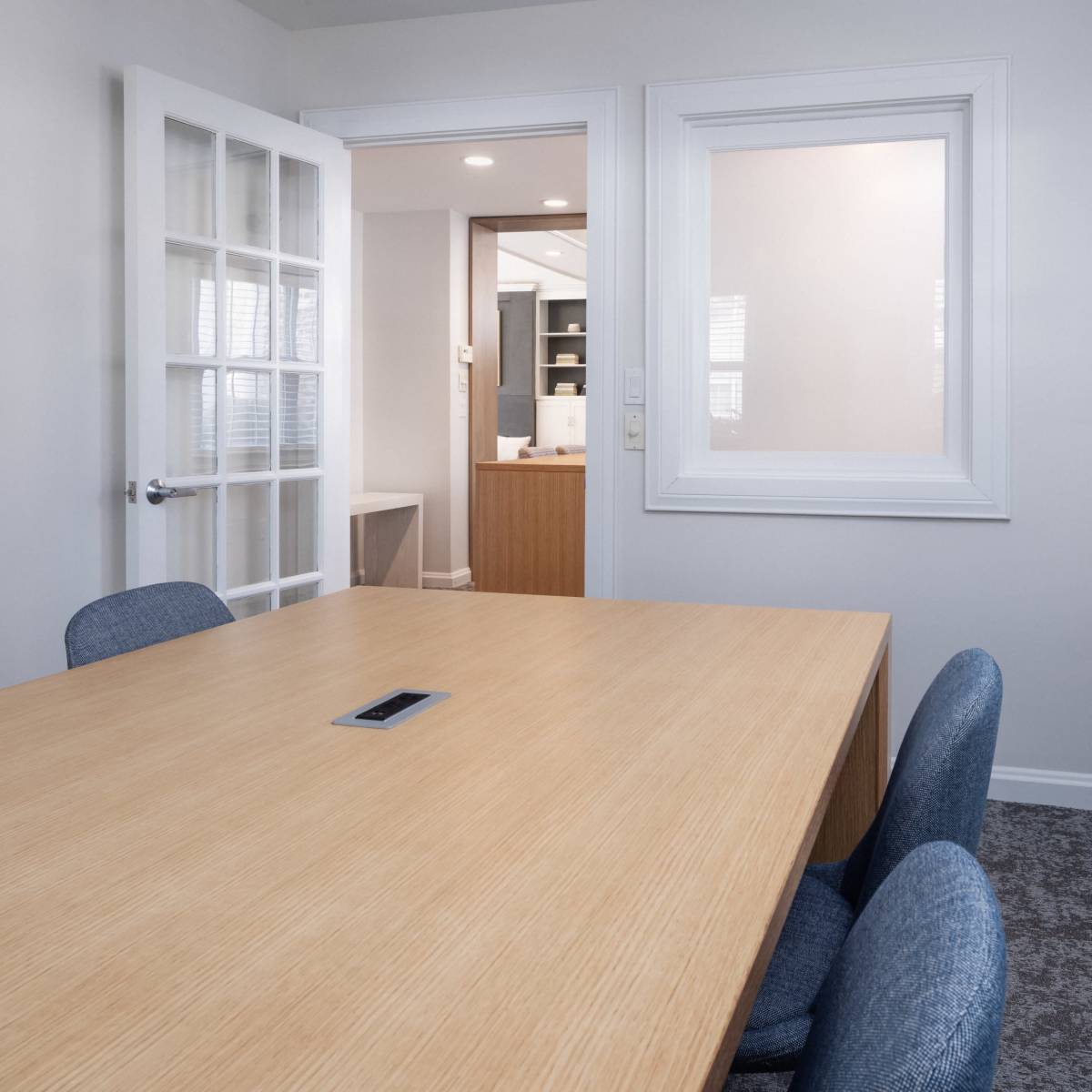 A conference room features a large wooden table surrounded by blue chairs, with a doorway leading to a nearby office space.