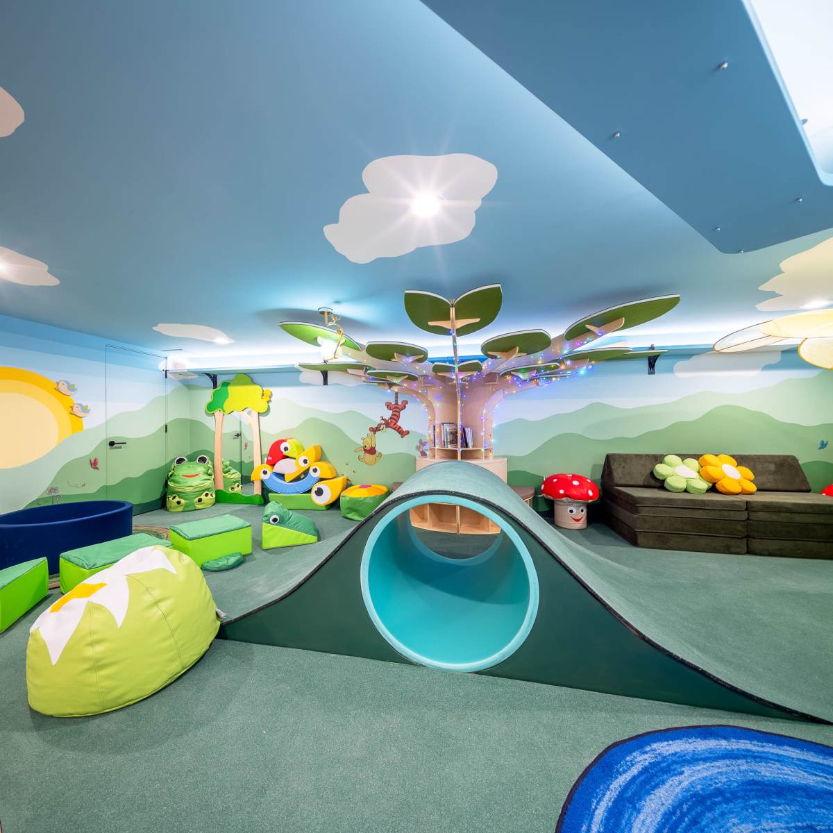 A colorful children's playroom features a slide, soft tunnels, and various playful decorations on the walls, including trees and clouds.