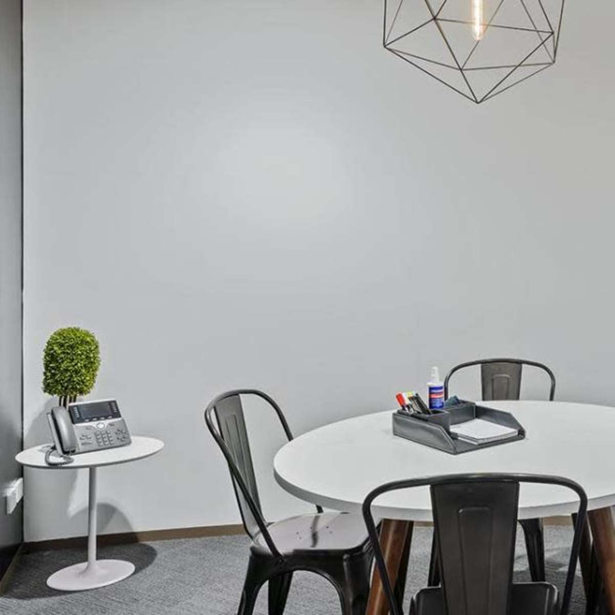 A round table surrounded by black chairs is set in a minimalist office space with a small plant and office supplies on the table.