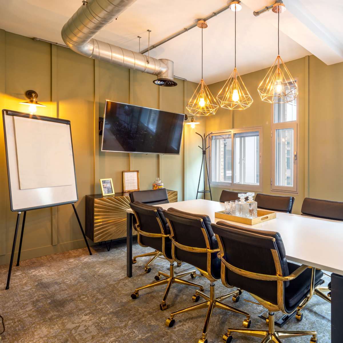 A modern conference room features a long table surrounded by black and gold chairs, a television mounted on the wall, and three pendant lights above.