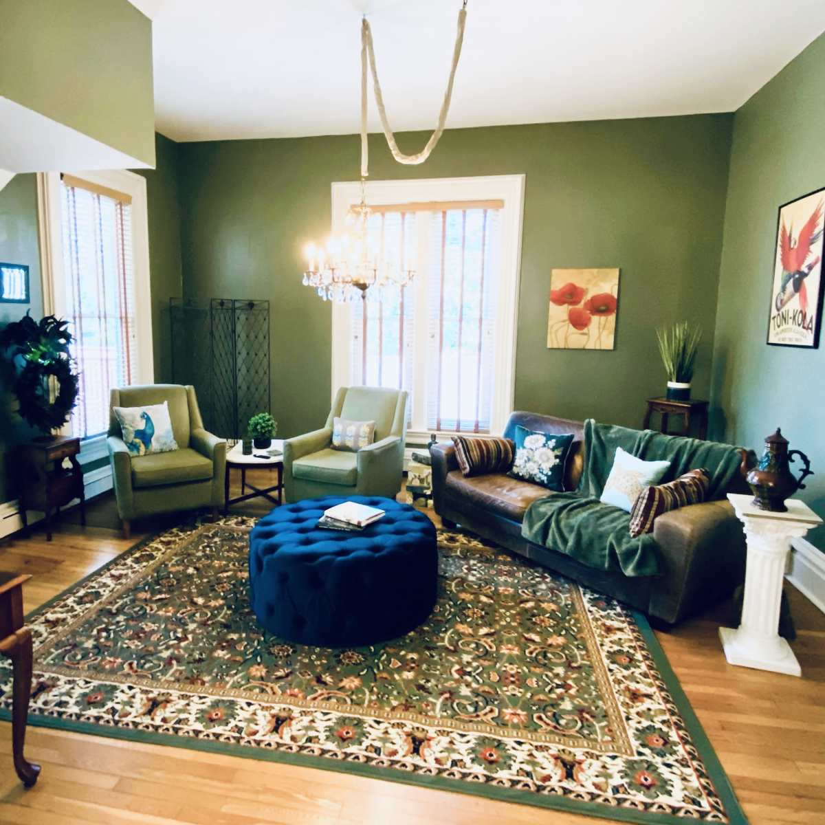 A cozy living room featuring a circular blue ottoman, a brown sofa with a throw blanket, two green chairs, and a decorative chandelier above, all set against green walls and a patterned area rug.