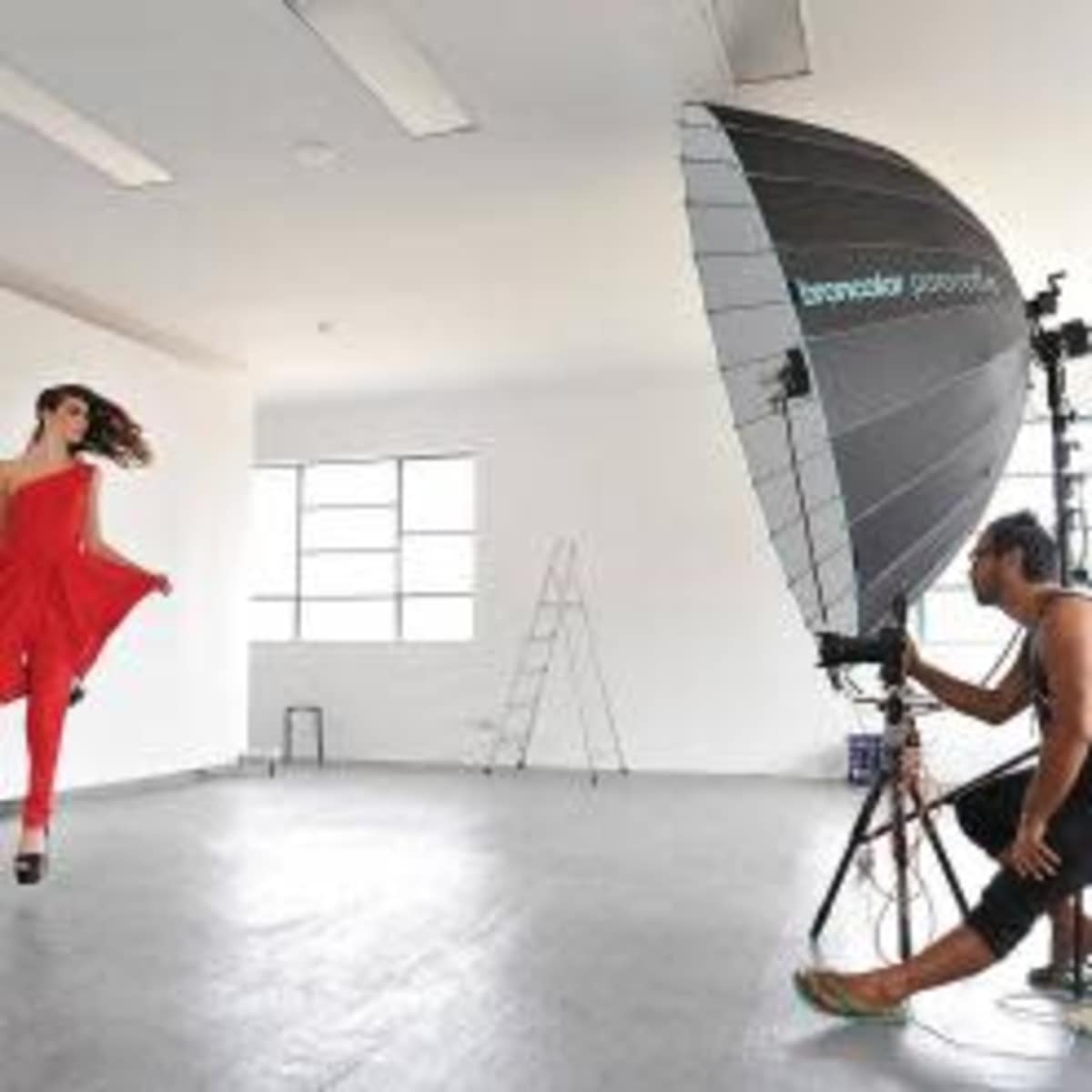 A model poses in a red outfit while a photographer adjusts lighting in a spacious studio.