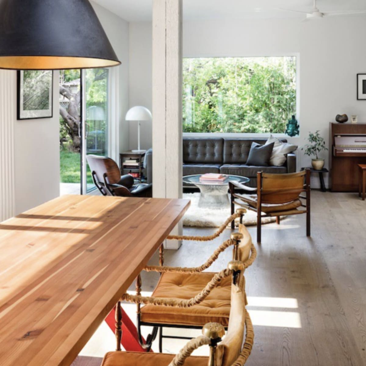 A wooden dining table with stylish chairs is positioned in a bright, open-plan living space featuring a large window that overlooks a verdant garden.