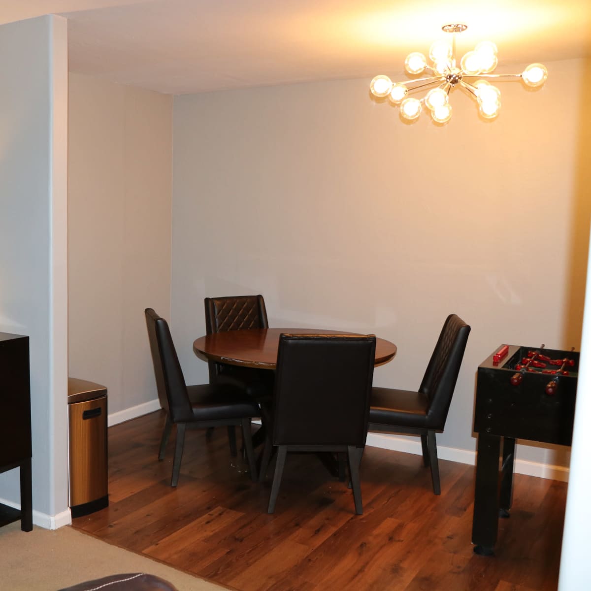 A modern dining area with a circular table and four chairs, adjacent to a dark wood sideboard and a lamp, set against walls of varying shades.