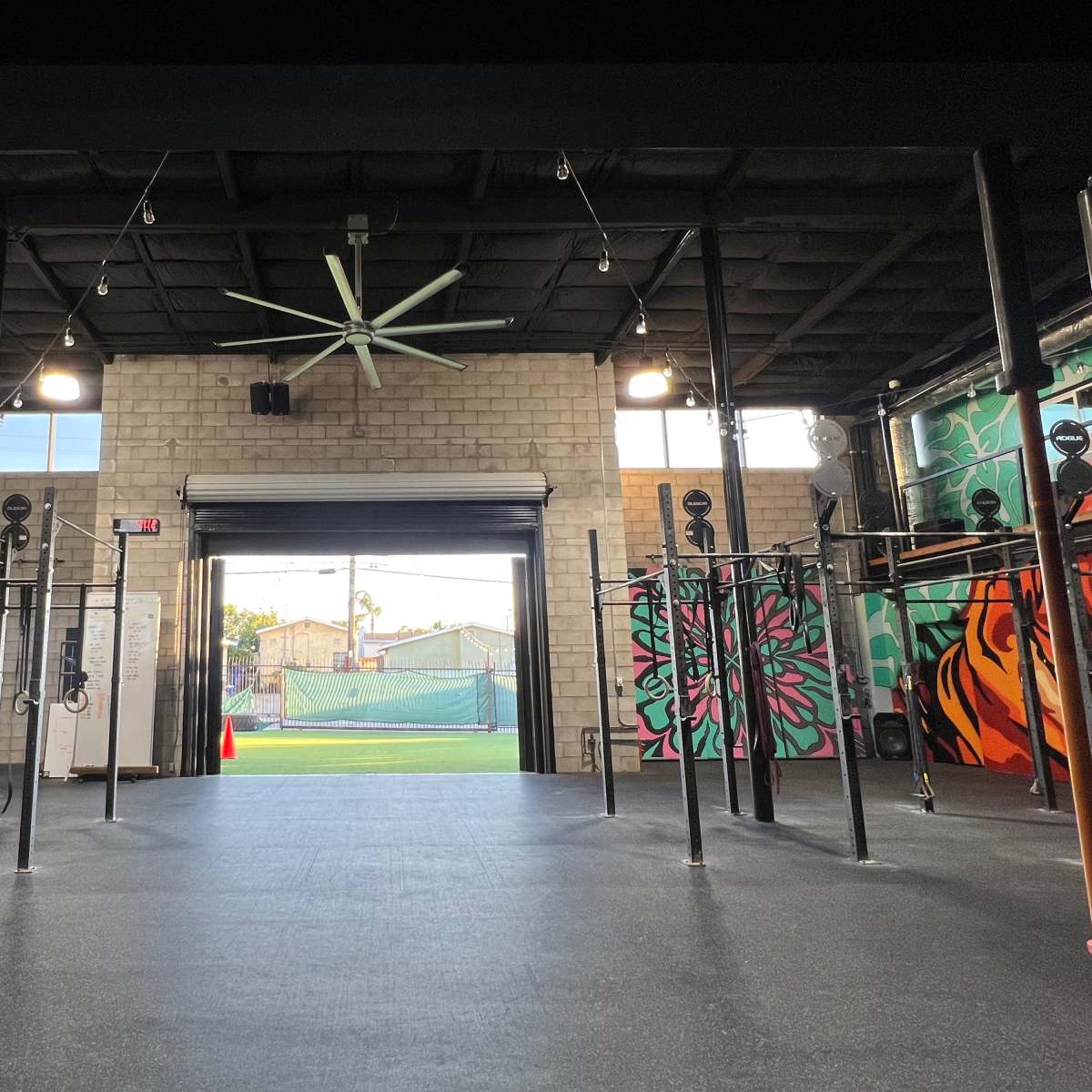 A gym interior with mounted exercise equipment and colorful murals on the walls, leading to an open area with a tennis court visible in the background.