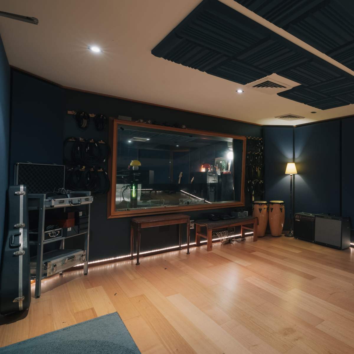 A recording studio with wooden flooring, blue soundproof walls, and a window revealing equipment in a separate room.