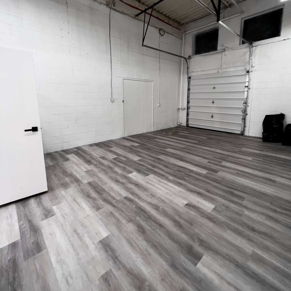 An empty industrial space with gray wooden flooring and a white wall with a door and a large overhead garage door.
