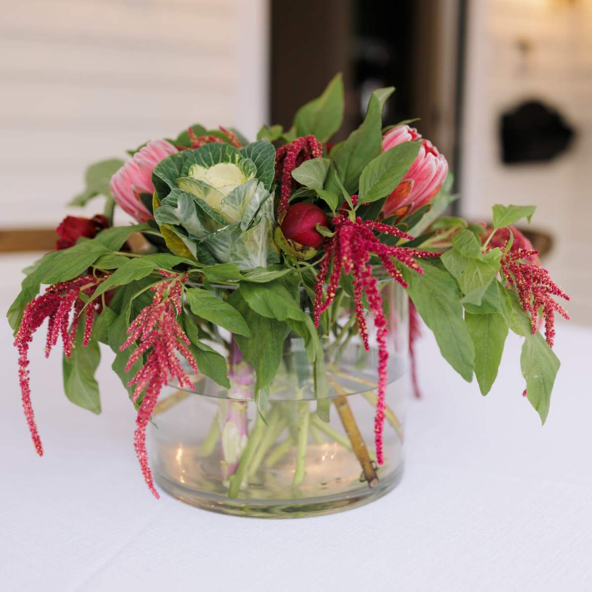 A clear glass vase holds a colorful arrangement of flowers, including roses, proteas, and green foliage, placed on a white tablecloth.