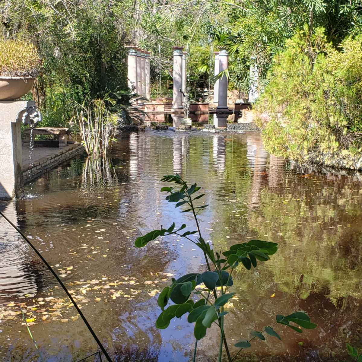 A calm pond is surrounded by lush greenery and features stone pillars in the background.