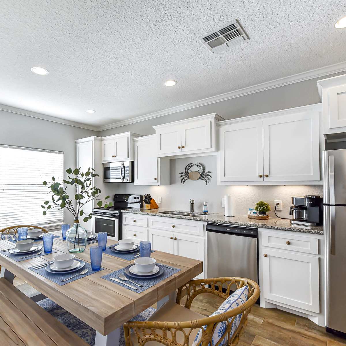 A modern kitchen features white cabinetry, stainless steel appliances, and a wooden dining table set for four with blue accents.