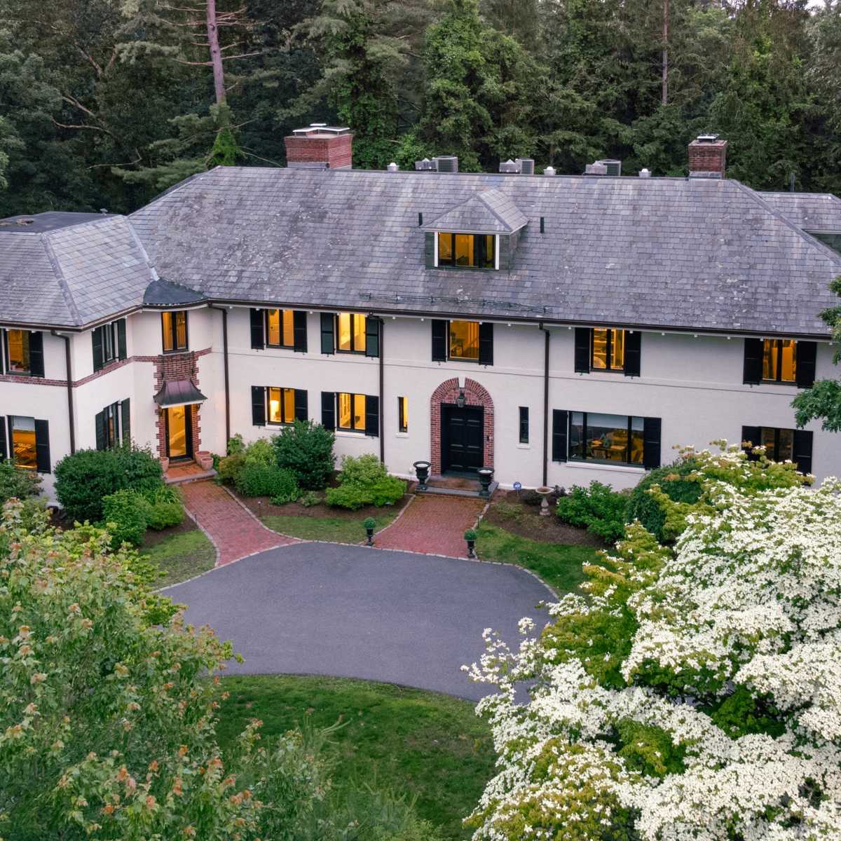 A large, multi-story house surrounded by trees and landscaping, with a winding driveway and large windows illuminated in the evening light.