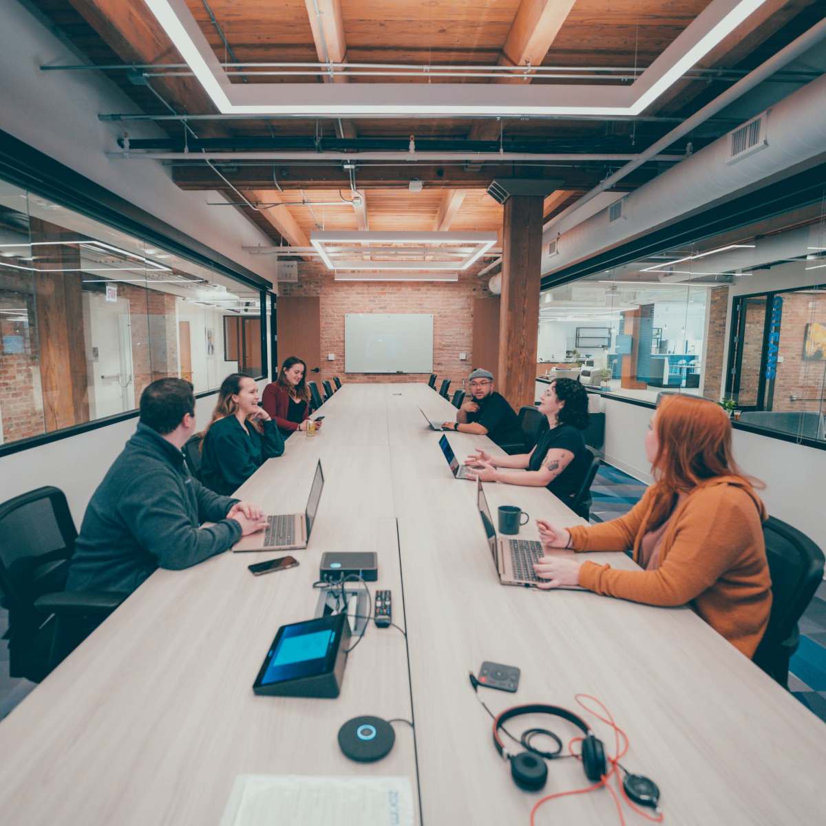 A group of eight people sits at a long conference table with laptops open, collaborating in a modern office space.