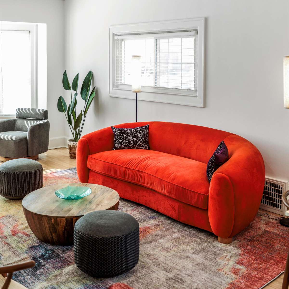 A bright living room features a curved orange sofa, a round wooden coffee table, and patterned ottomans, with natural light streaming through a window.