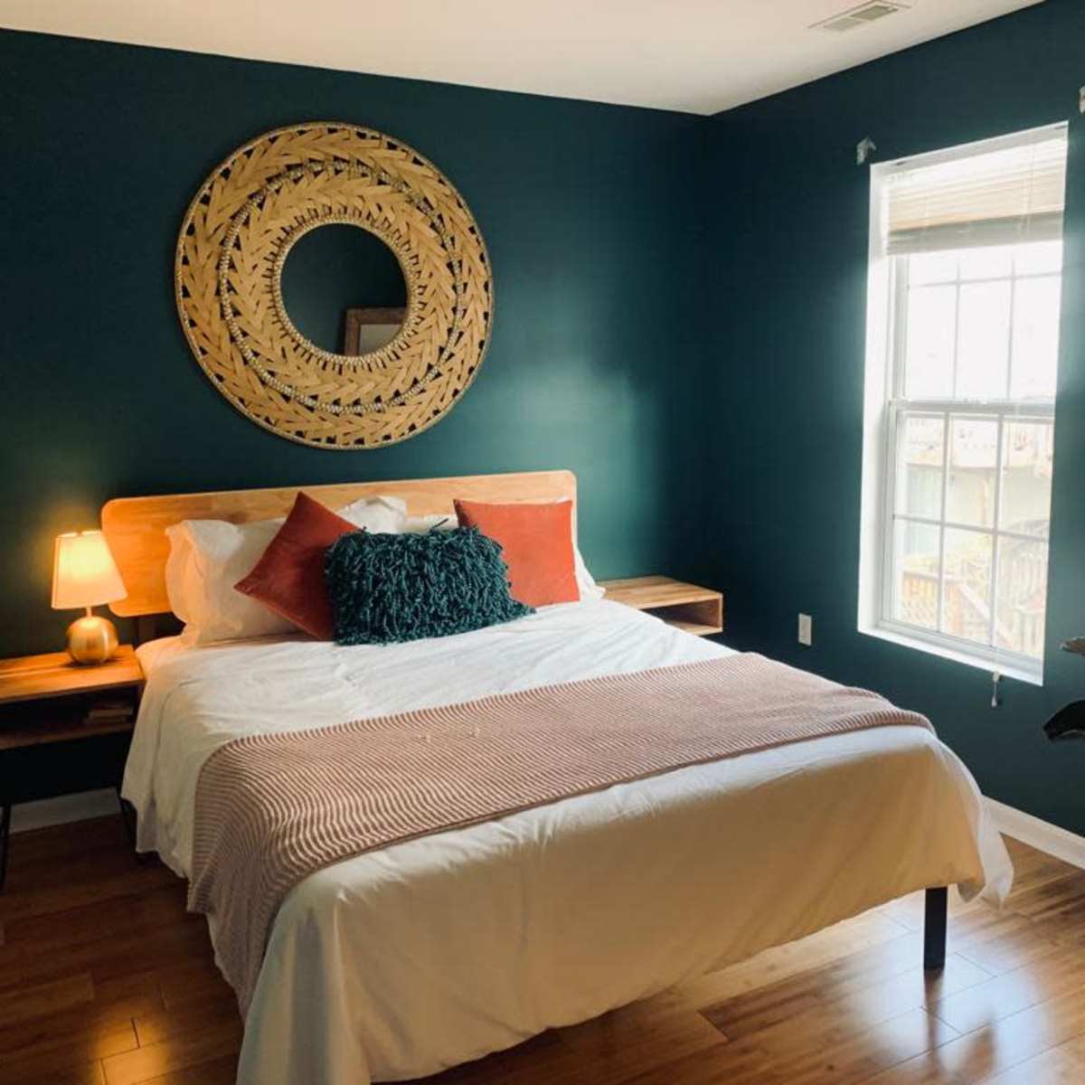 A neatly arranged bedroom featuring a bed with white linens, orange and teal pillows, a round decorative mirror on the wall, and a window allowing natural light.