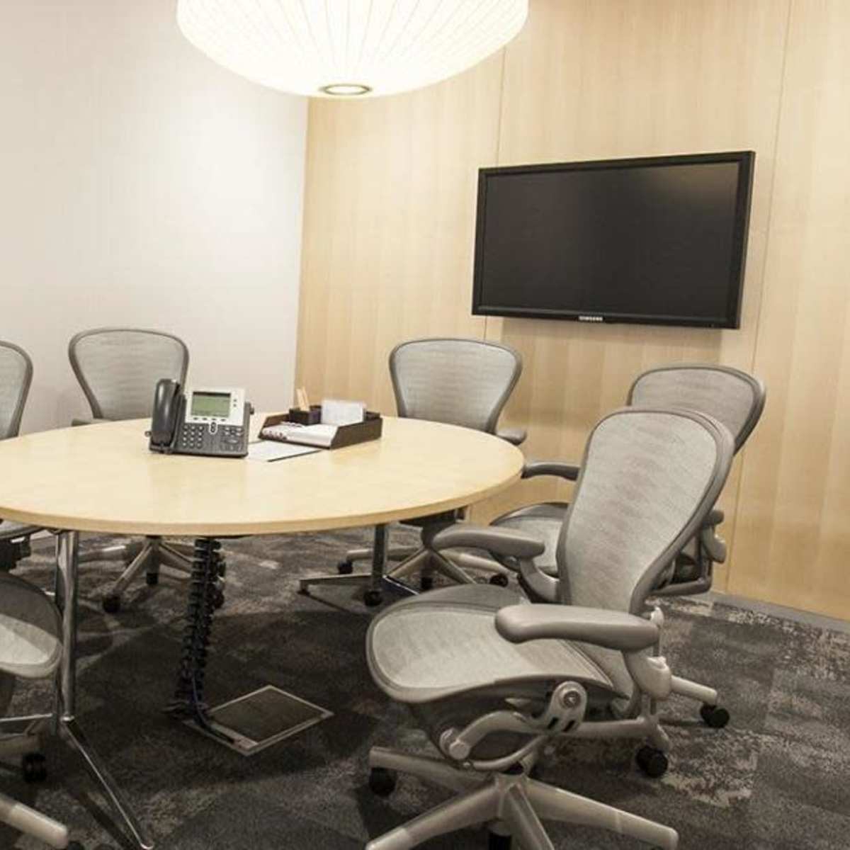 A conference room features a round table surrounded by six ergonomic chairs, a telephone, and a wall-mounted screen.