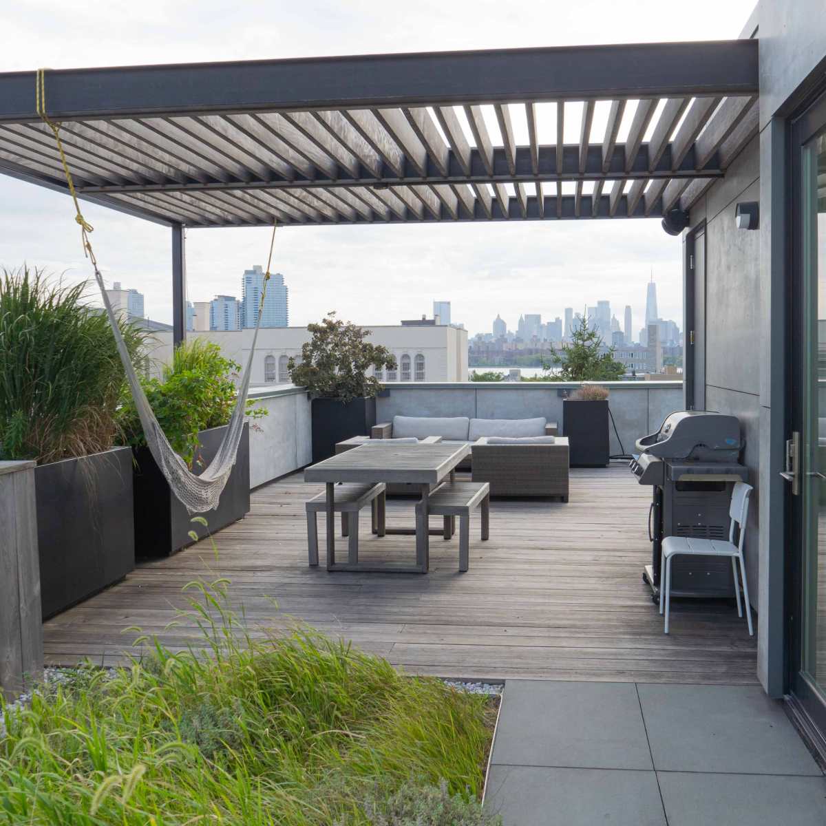 A rooftop terrace with wooden decking, a dining table, a hammock, lounge seating, and a skyline view in the background.