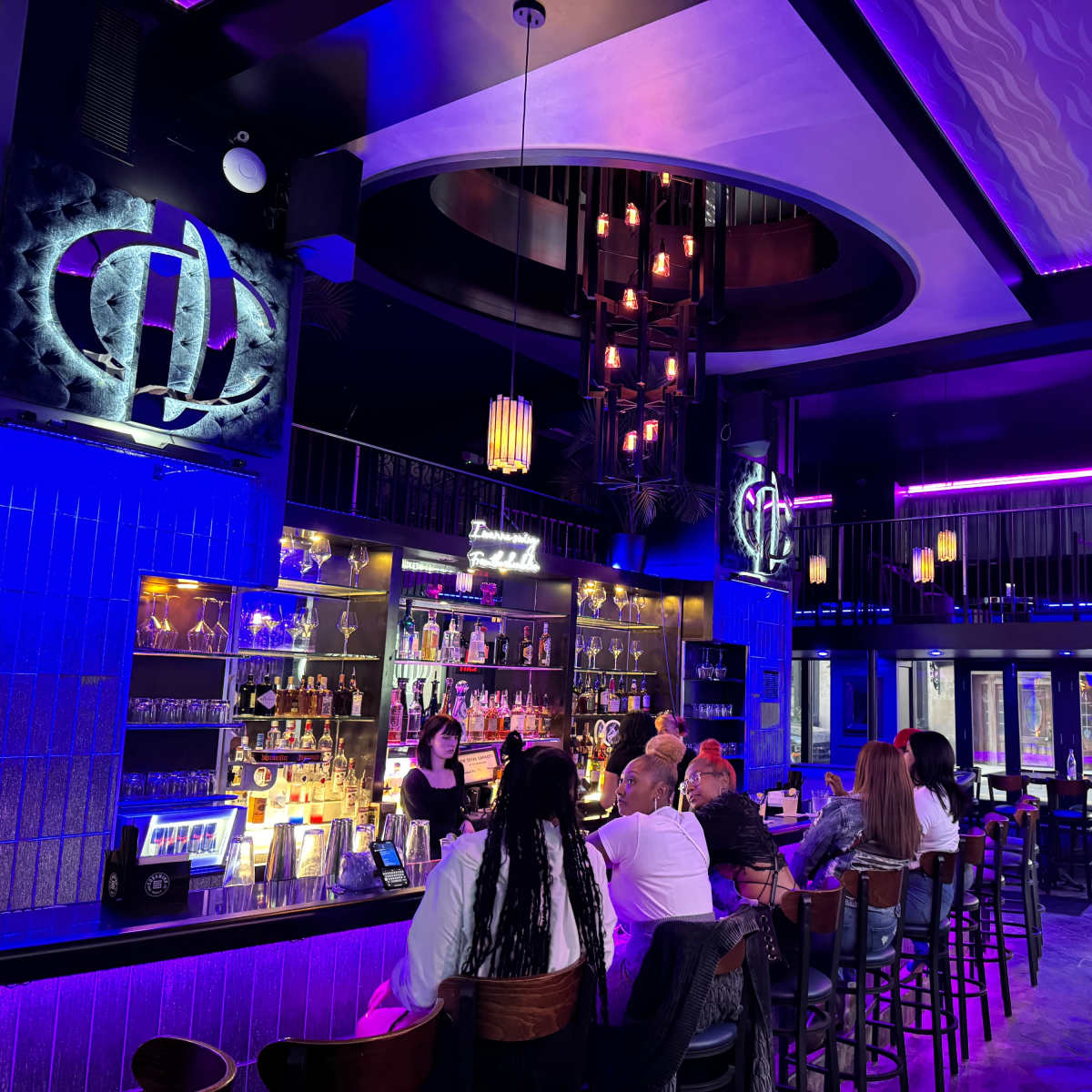A vibrant bar interior with purple lighting, a long counter filled with bottles, and patrons seated at tables and the bar.