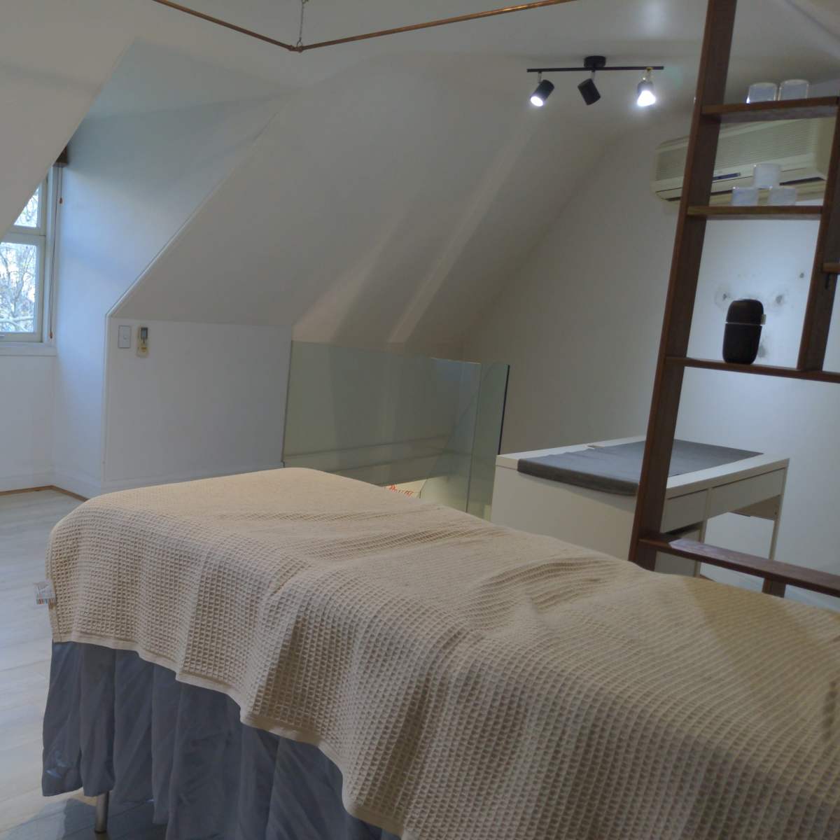 A clean, well-lit room with a treatment table covered in a beige cloth, a stool, and a work surface against a slanted wall.