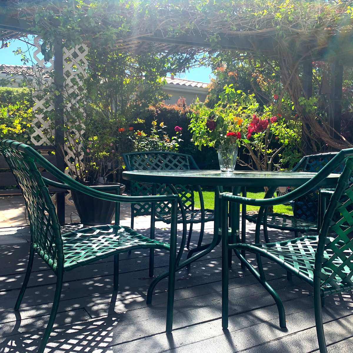 A shaded patio features a round table surrounded by four green metal chairs, with colorful flowers and greenery in the background.