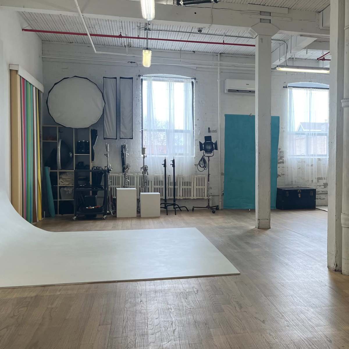 A spacious, well-lit studio with wooden floors, large windows, and various photography equipment arranged along the walls.