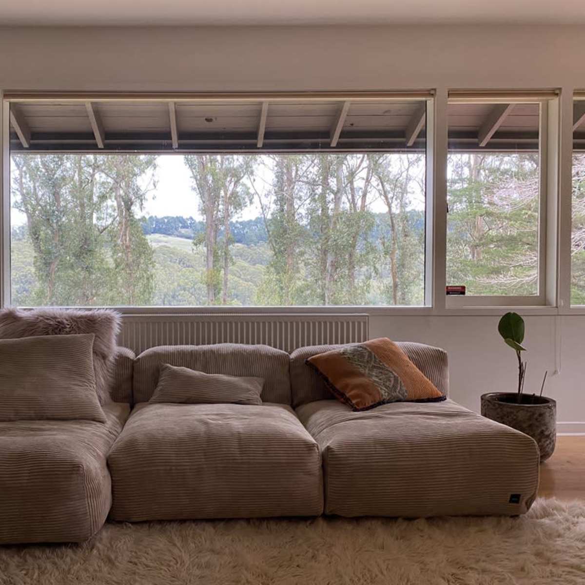 A large, upholstered sectional sofa is positioned in front of wide windows that offer a view of lush green hills and trees.