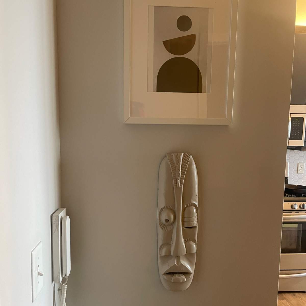 A white wall features a framed abstract artwork above a carved white mask, next to a wall-mounted telephone.