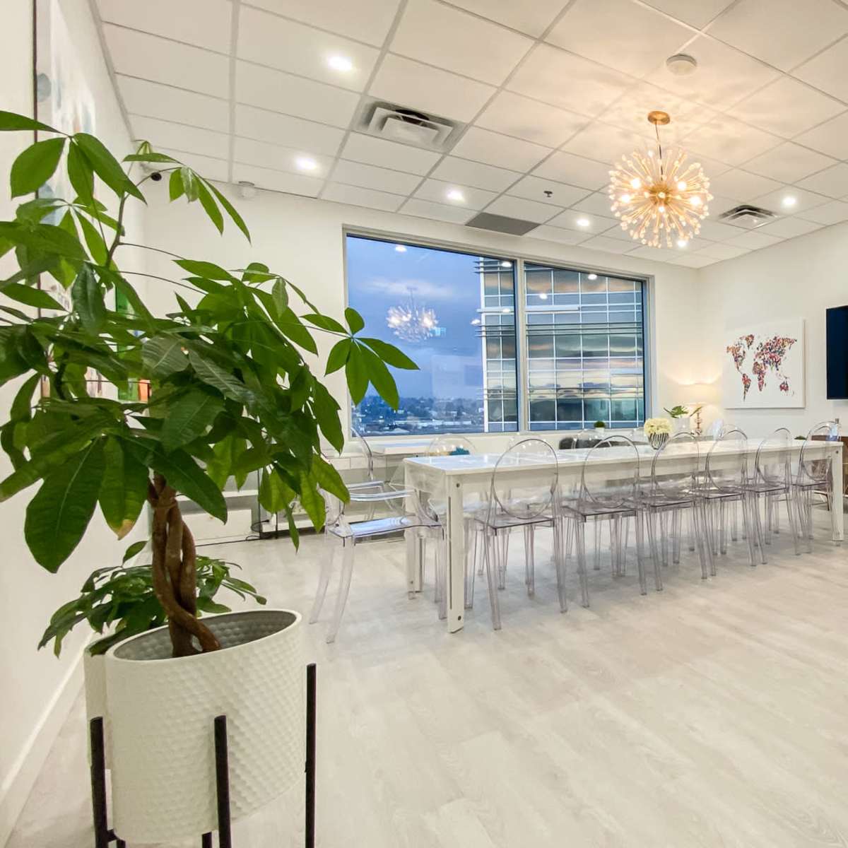 A modern conference room with a long table, transparent chairs, a large TV, and a view of the city through a window.