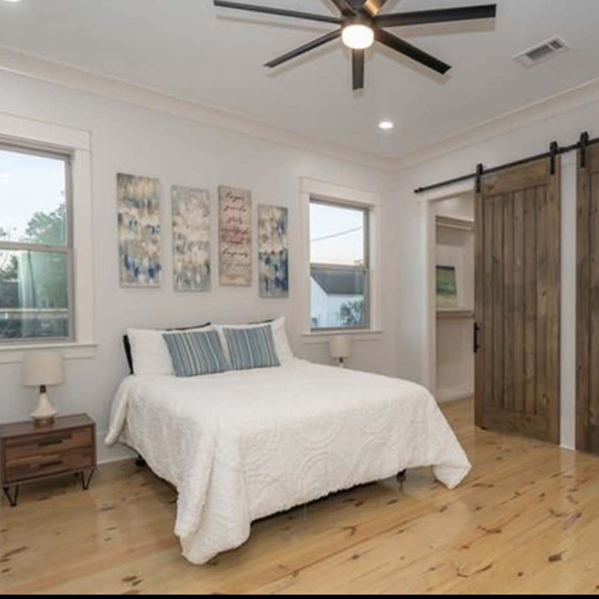 A modern bedroom with a white quilted bedspread, wooden floors, and sliding barn doors leading to a bathroom.