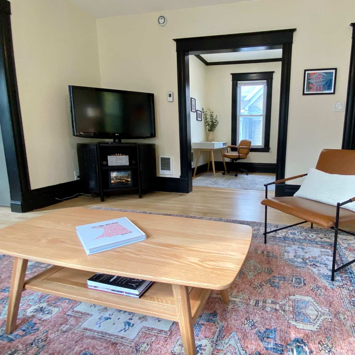 The living room features a wooden coffee table, a leather armchair, a television, and a desk in a bright, organized space.