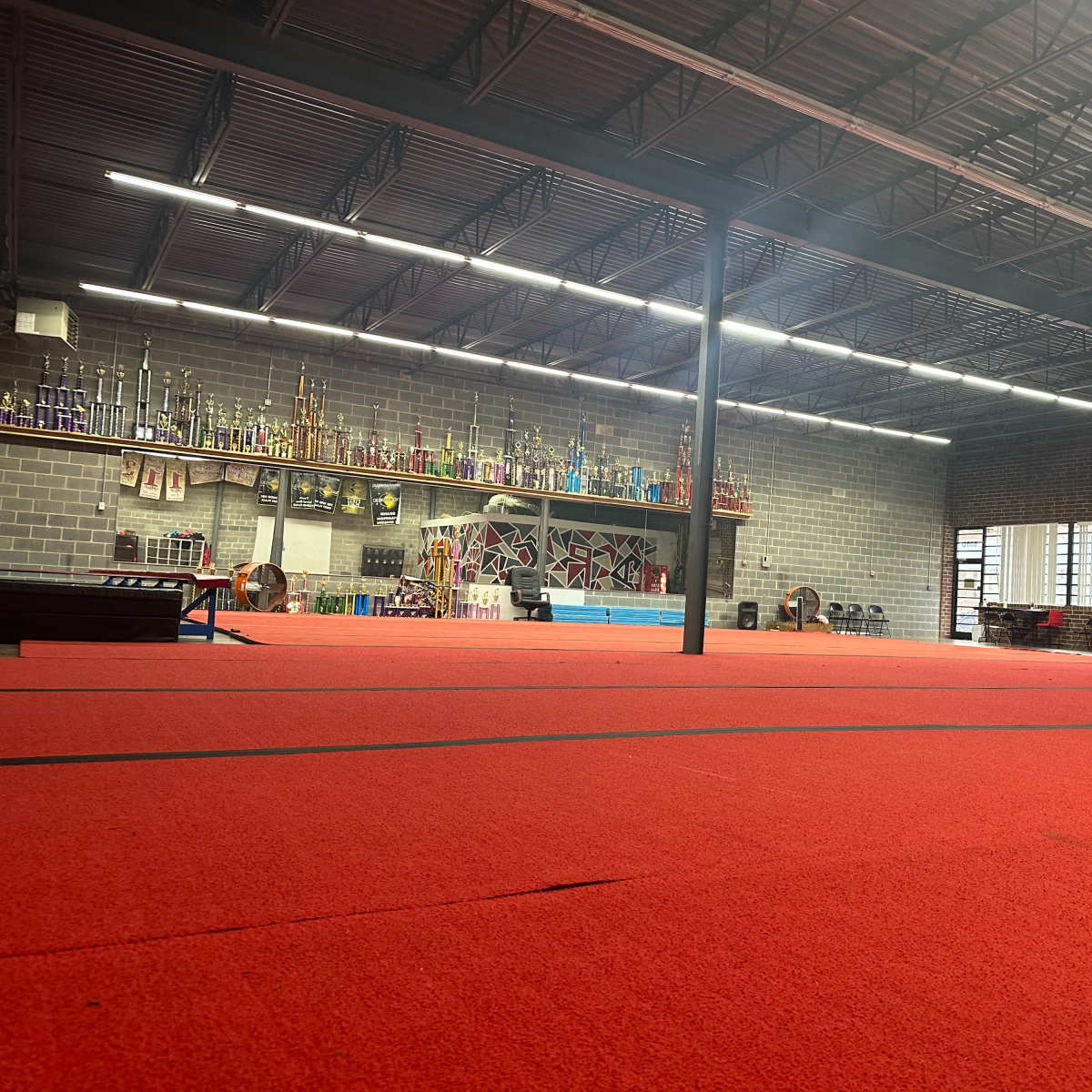 A gymnasium with red flooring, trophy displays along the back wall, and various gym equipment arranged throughout the space.