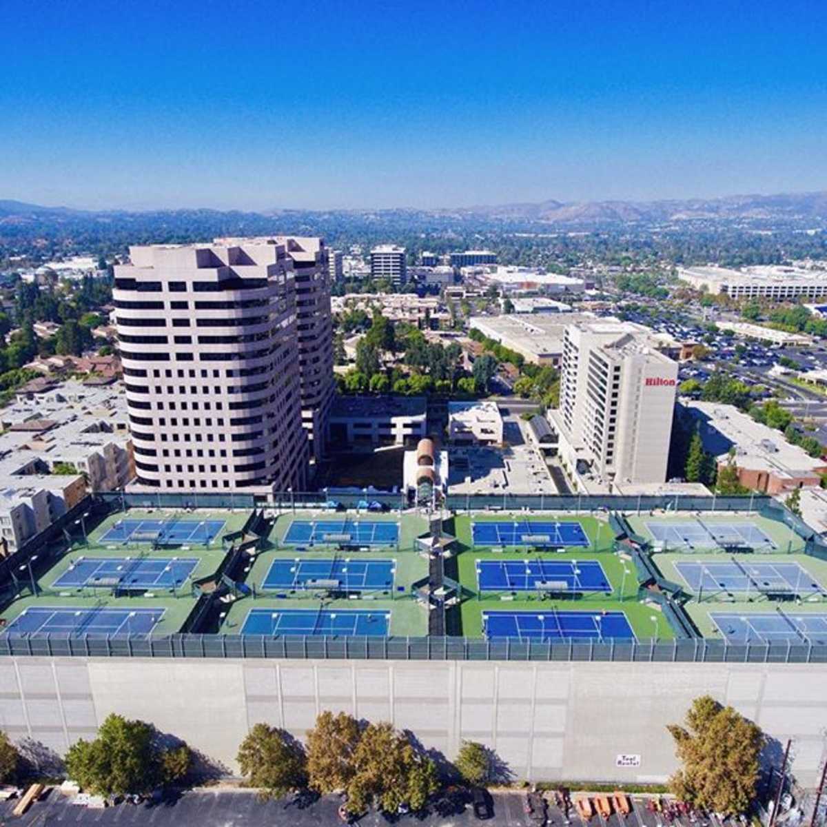 A rooftop tennis facility with multiple courts surrounded by urban buildings and a clear blue sky.