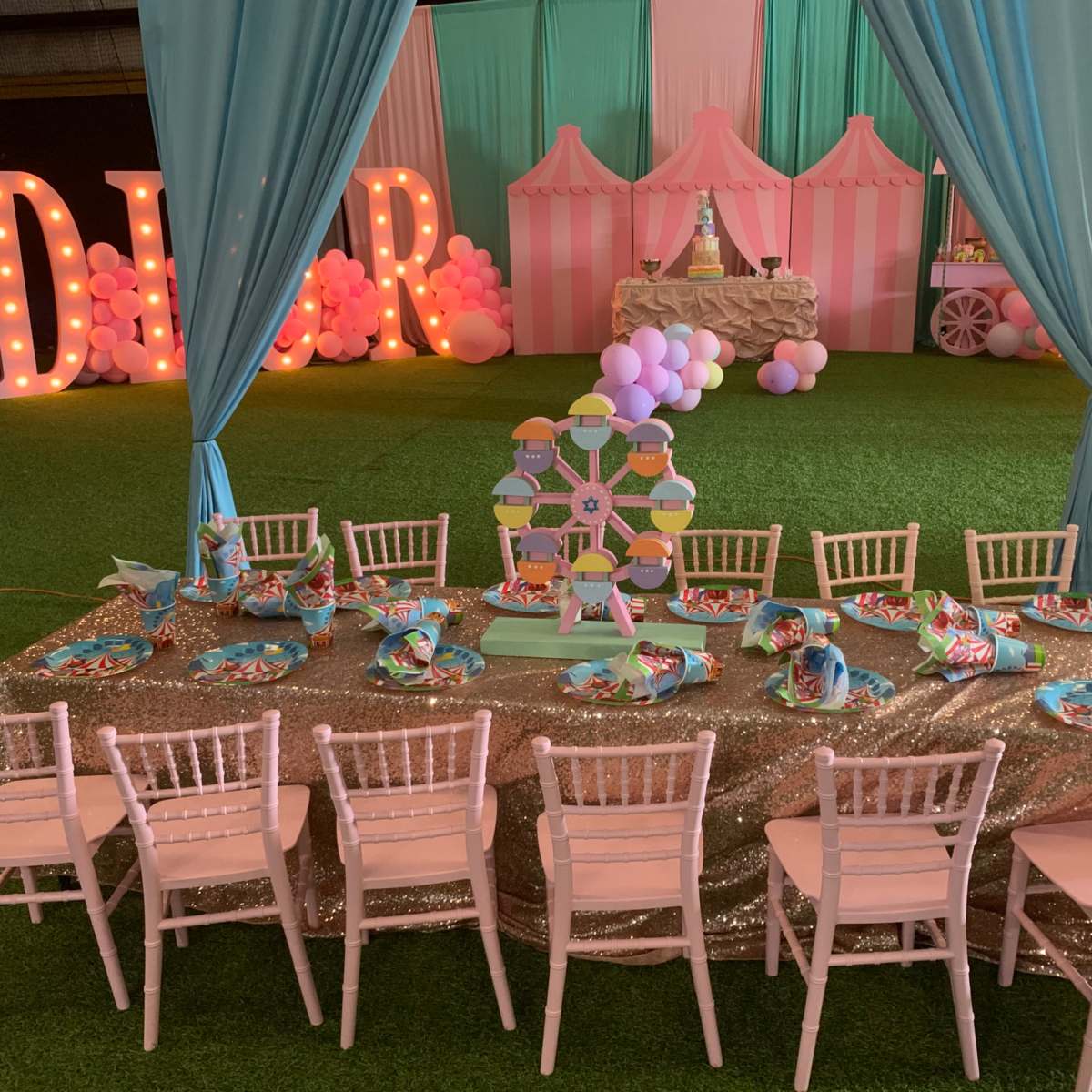 A decorated event space features a long table set with colorful party plates and chairs, surrounded by pastel drapes and festive decorations.
