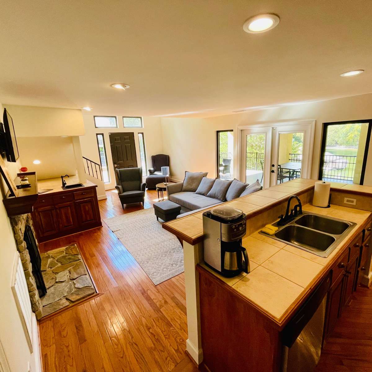 A spacious living area with a kitchen bar, featuring a gray sofa set, a stone fireplace, and large windows overlooking an outdoor patio.