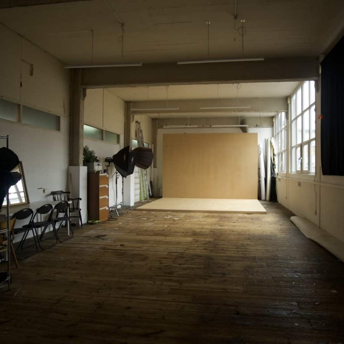 A spacious interior with wooden floors, large windows, and a blank backdrop set against a wall, ideal for photography or art projects.