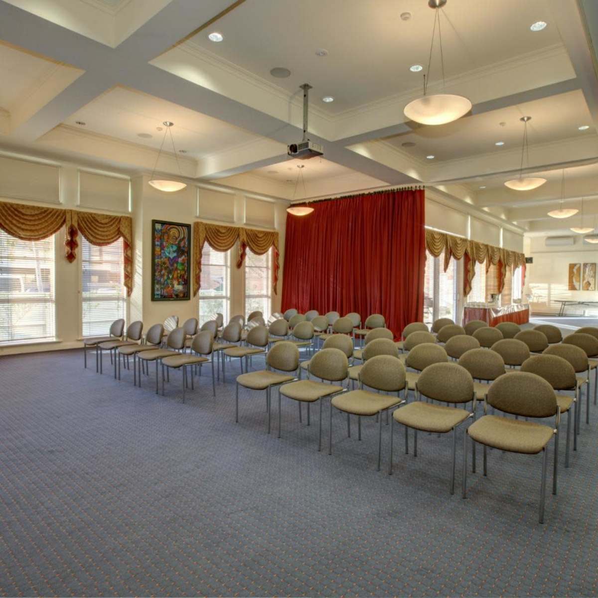 A meeting room set up with rows of circular chairs facing a red curtain, with large windows and decorative drapes along the walls.