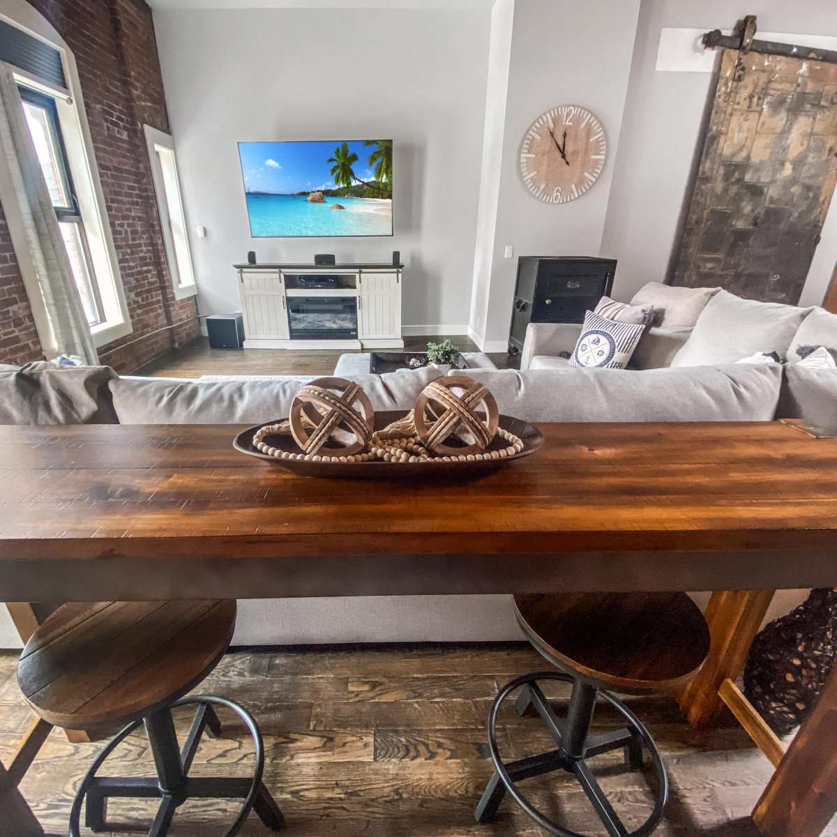 A rustic wooden table with two stools is placed in front of a living room featuring a television, a beach scene on the screen, and an exposed brick wall.