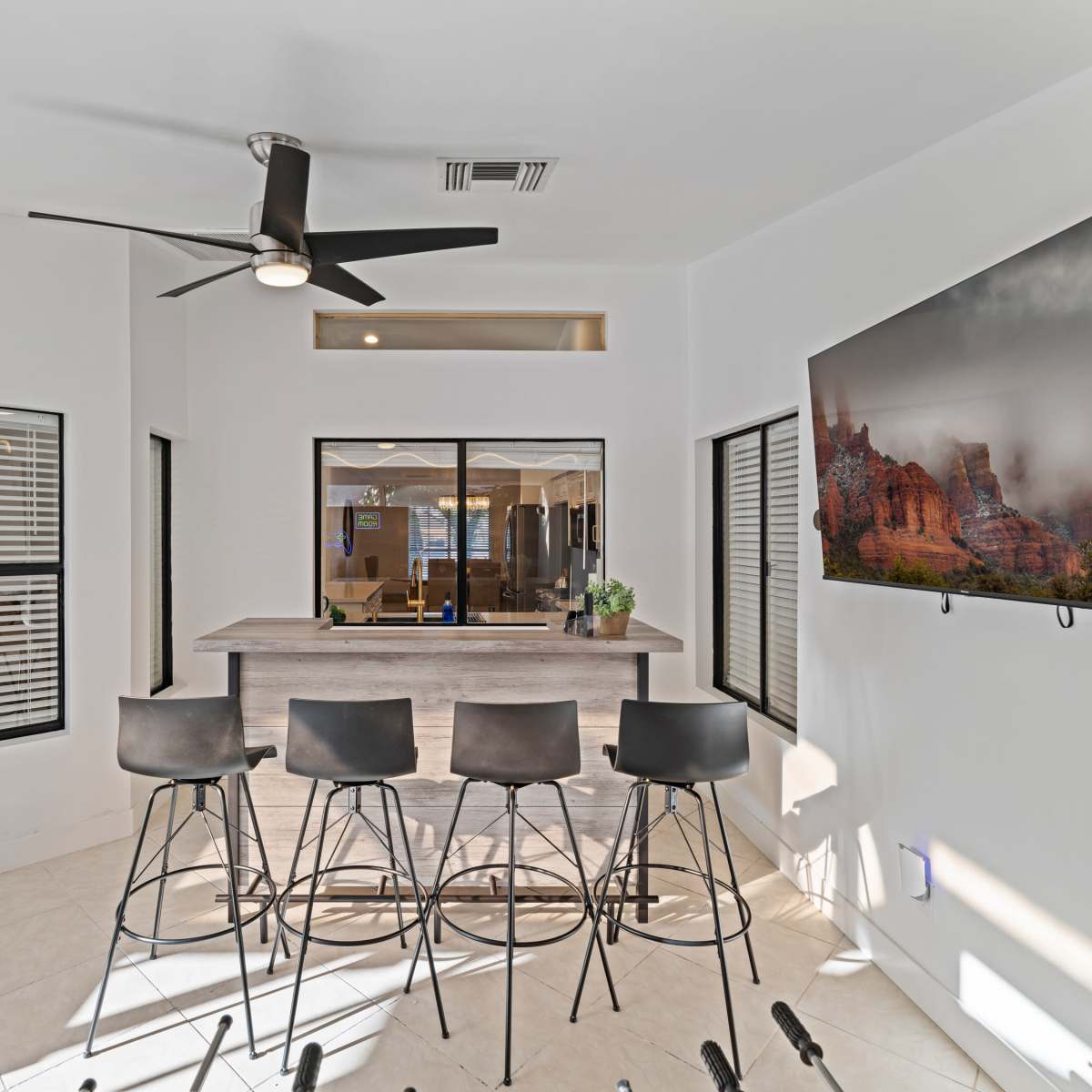 A modern indoor bar area with high stools, a ceiling fan, large windows, and a mounted TV on the wall.