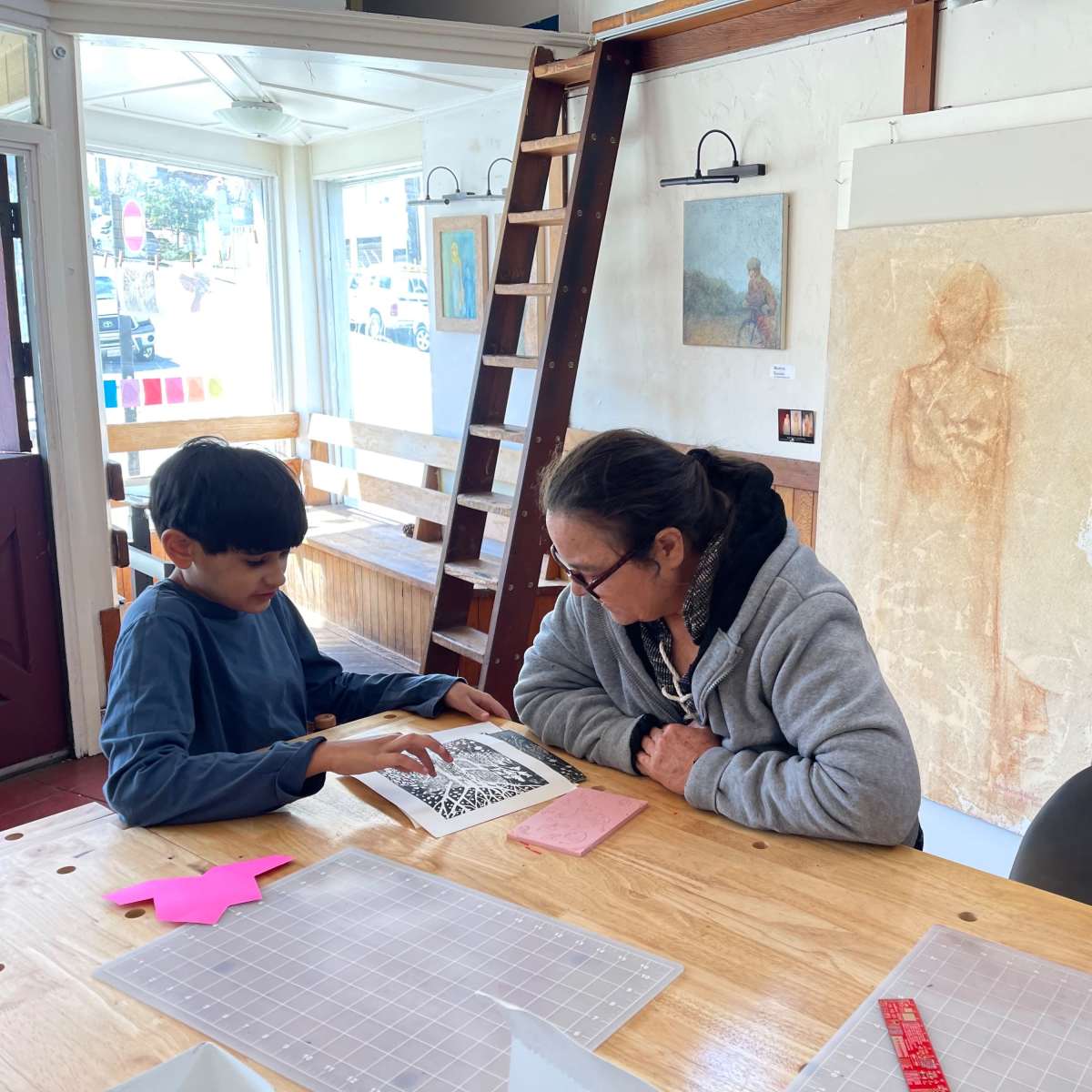 A child and an adult sit at a wooden table, examining a book, while artwork hangs on the walls of a well-lit space.