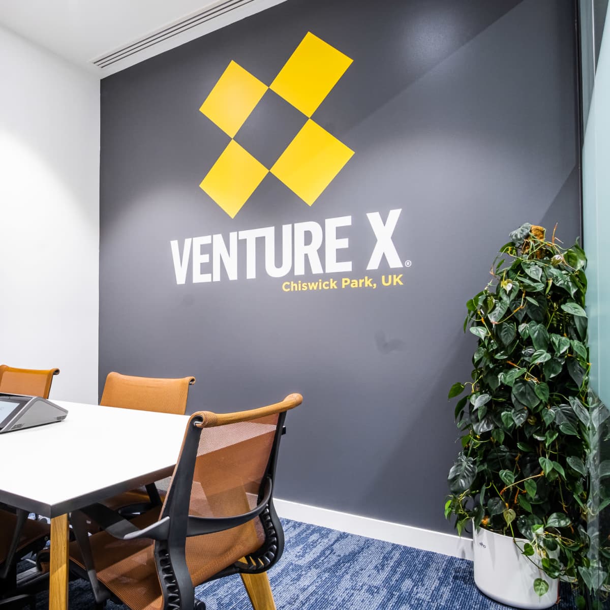 A modern conference room featuring a table, chairs, a wall-mounted screen, and a large logo of "Venture X" alongside a green plant.