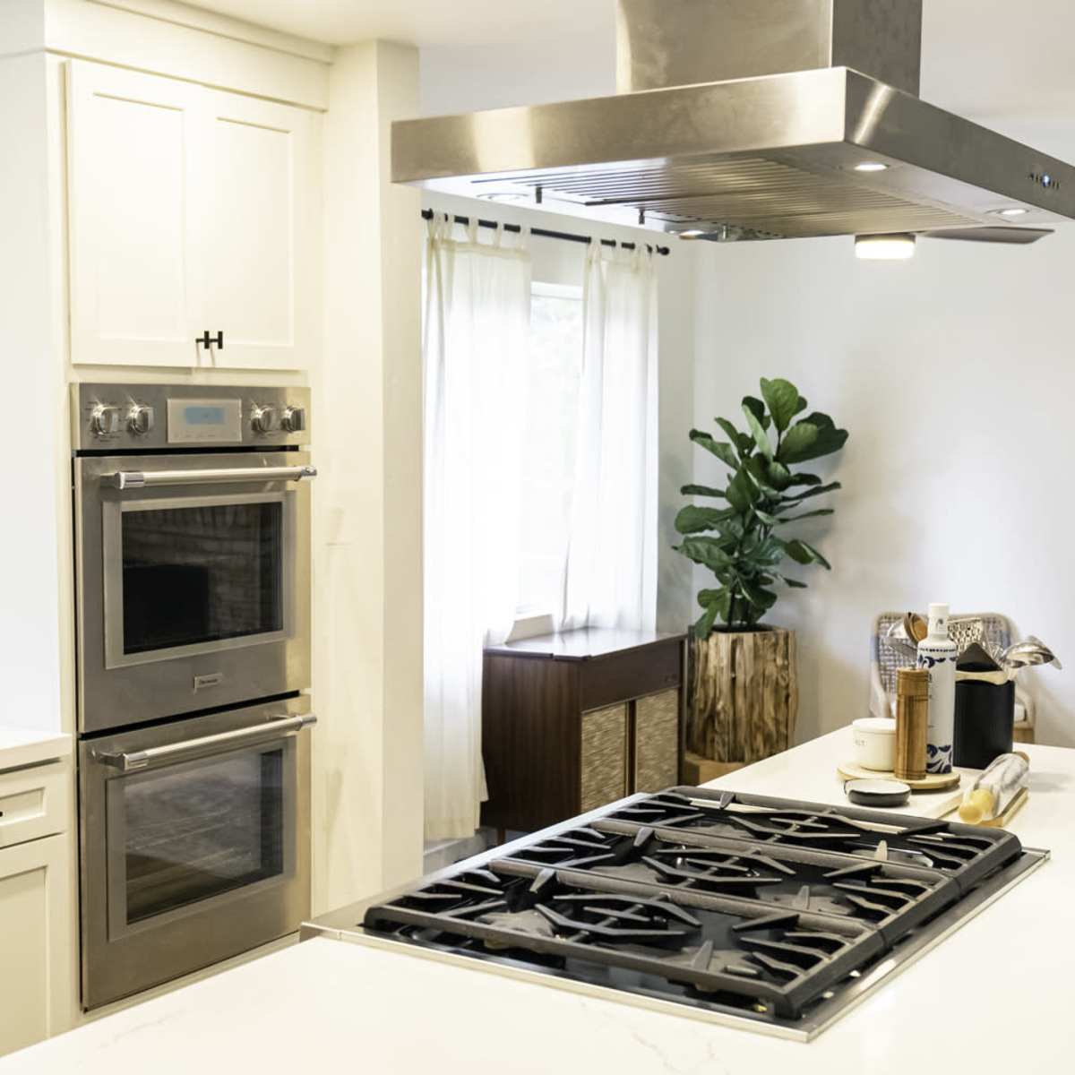 A modern kitchen featuring a gas cooktop, double wall ovens, a stainless steel range hood, and a window with sheer curtains.
