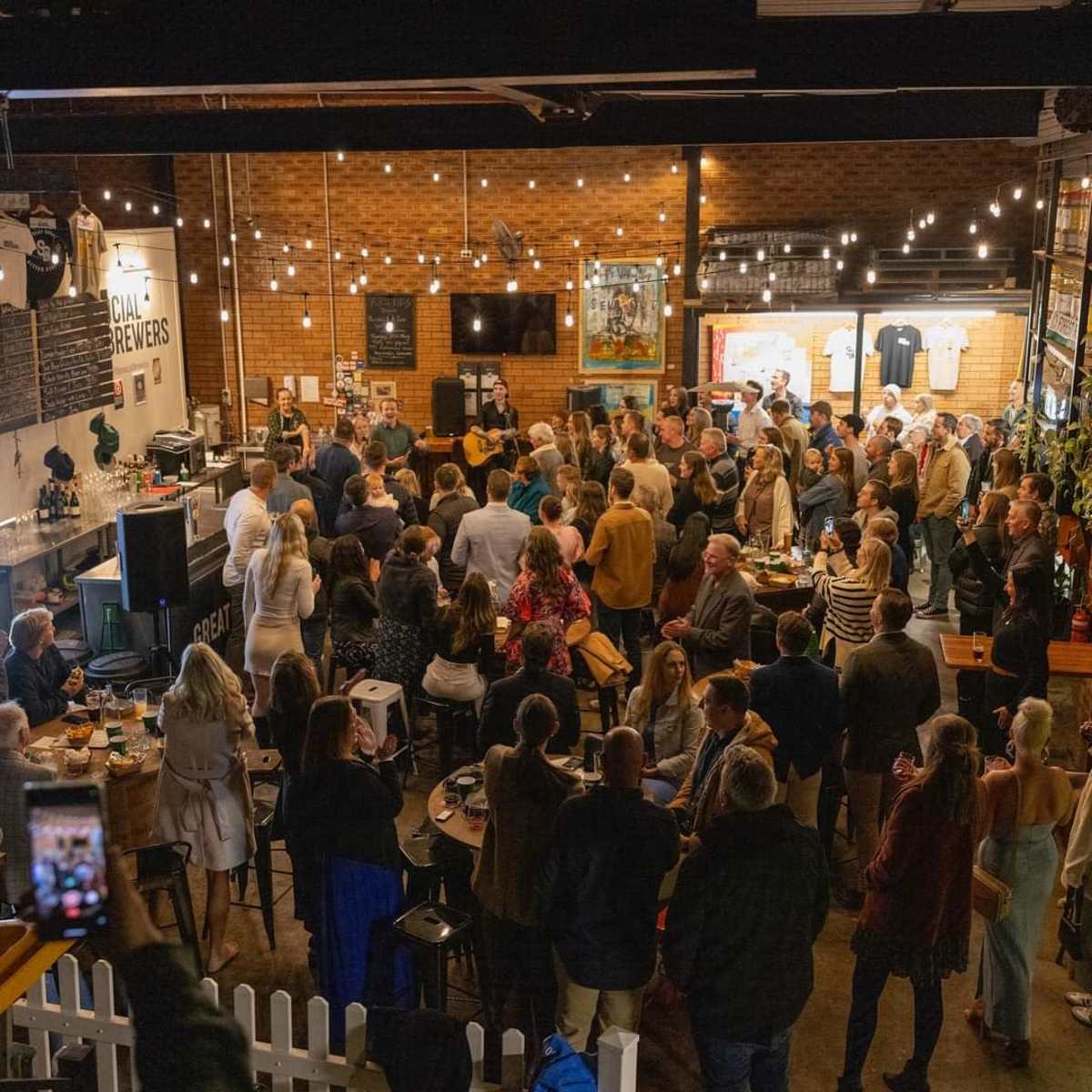 A large crowd gathers in a brewery, with people socializing and drinks on tables under string lights.