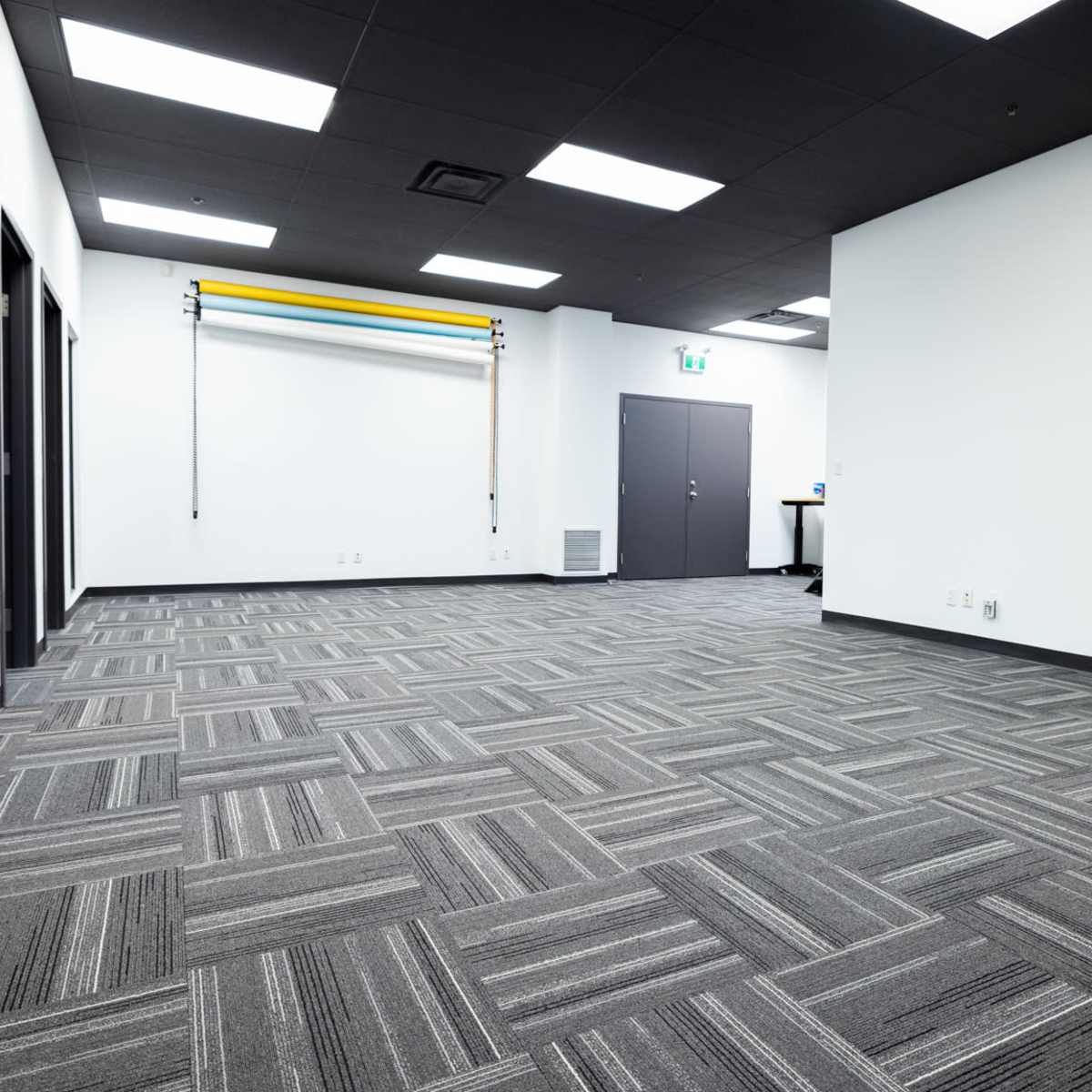 A large, empty room with gray carpeting, white walls, and overhead fluorescent lighting, featuring mirrors and a closed door on one side.