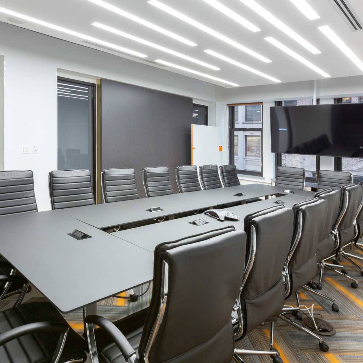 A modern conference room features a long black table surrounded by multiple black ergonomic chairs, with a large television mounted on the wall and illuminated by overhead LED lights.